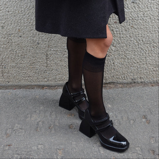 Person wearing black shoes and a dark coat with a quilted handbag on a gray pavement.