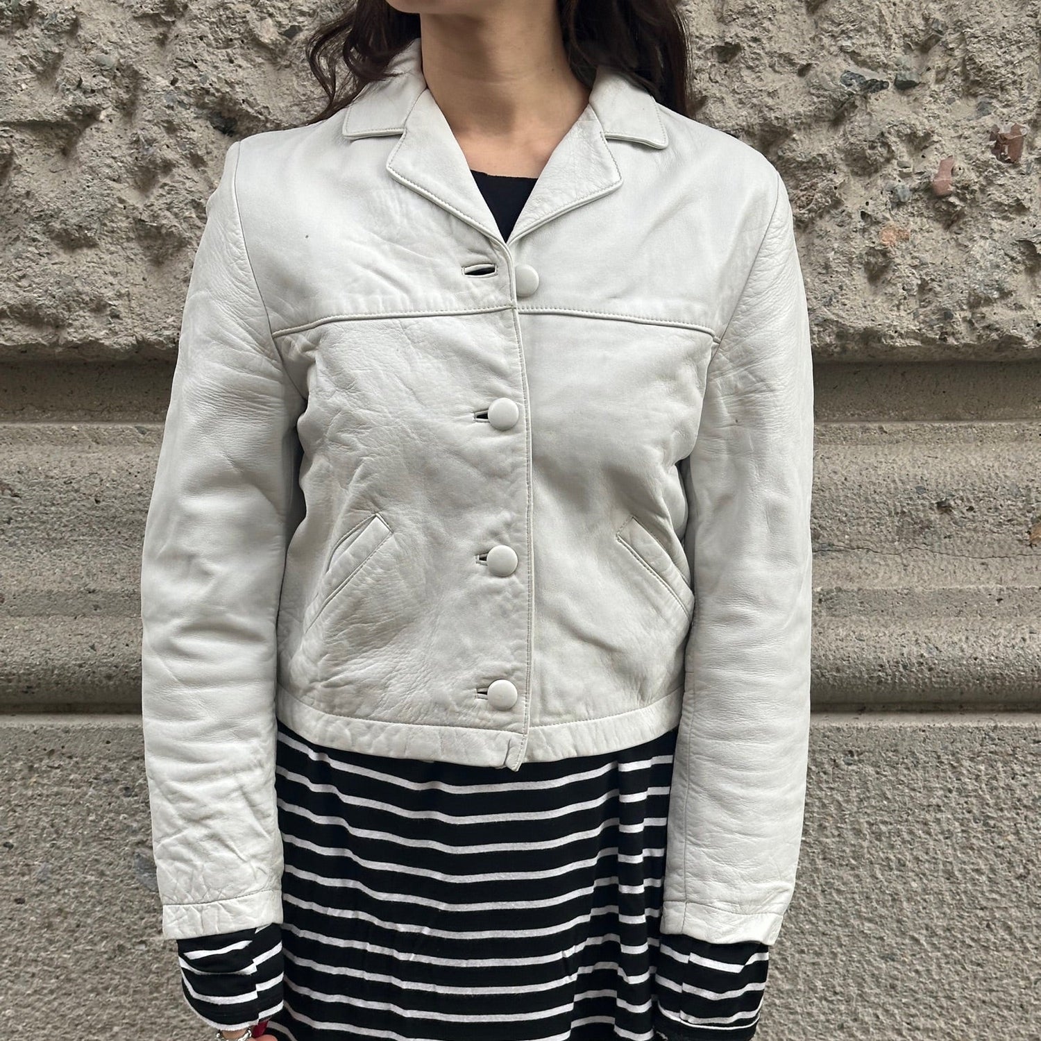 Person wearing a light-colored jacket and black and white striped skirt against a stone wall.