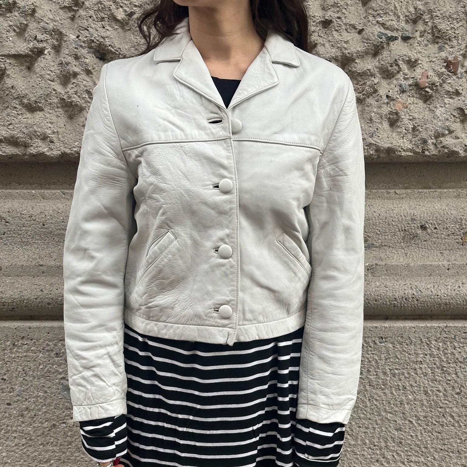 Person wearing a light-colored jacket and black and white striped skirt against a stone wall.