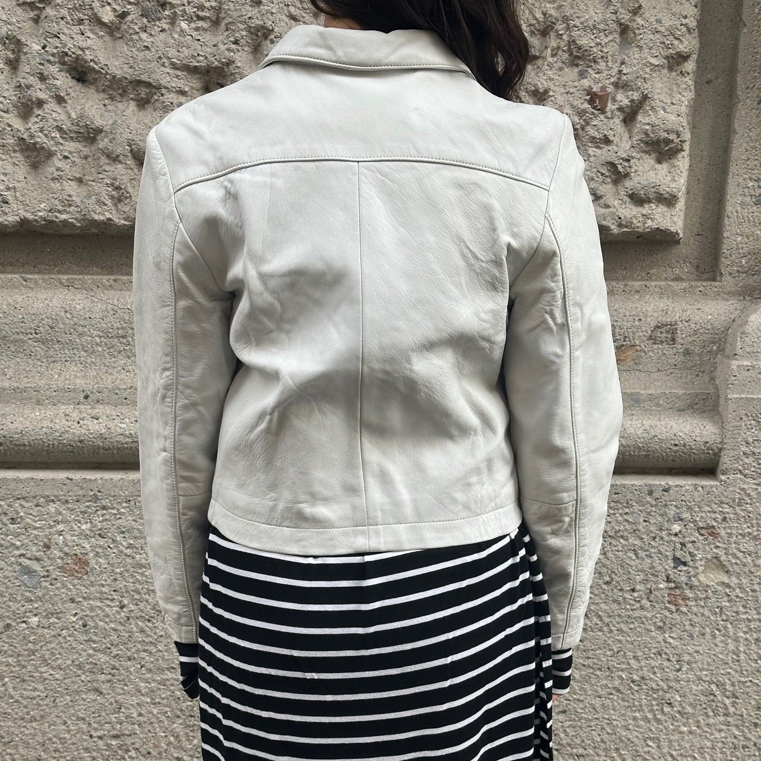 Person wearing a light-colored jacket and black and white striped skirt against a stone wall.