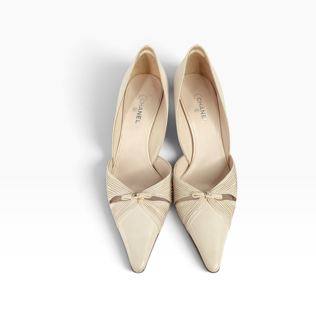 A pair of Chanel pointed-toe pumps in a light beige/cream color, featuring a textured fabric upper and a small satin bow detail on the toe box, photographed from a top-down angle on a white background.