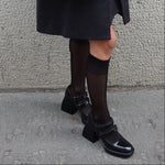 Person wearing black shoes and a dark coat with a quilted handbag on a gray pavement.