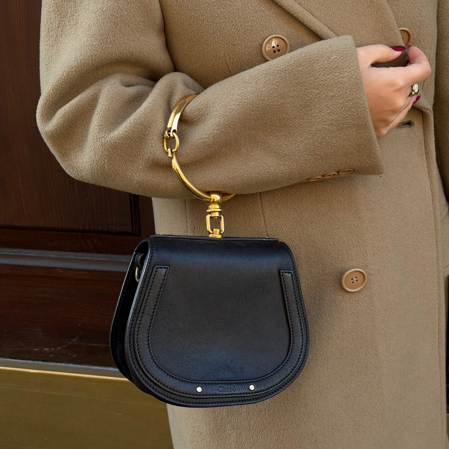 Person wearing a beige coat holding a black handbag with gold accents.