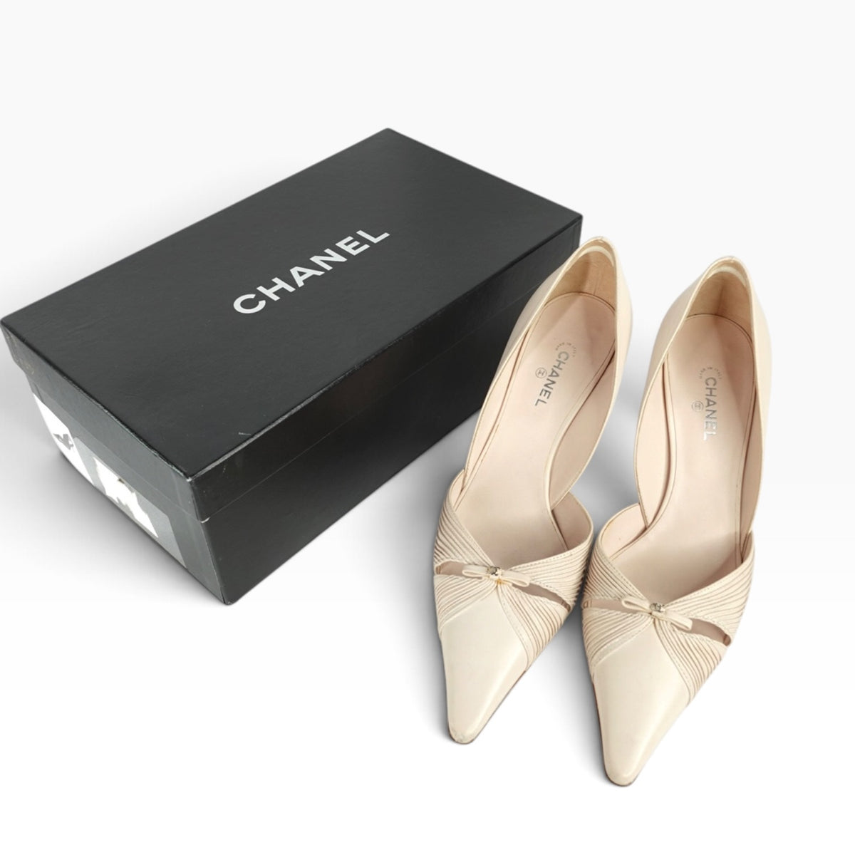 A pair of Chanel pointed-toe pumps in a light beige/nude color with a small bow detail at the instep, displayed next to their black Chanel shoebox on a clean white background.