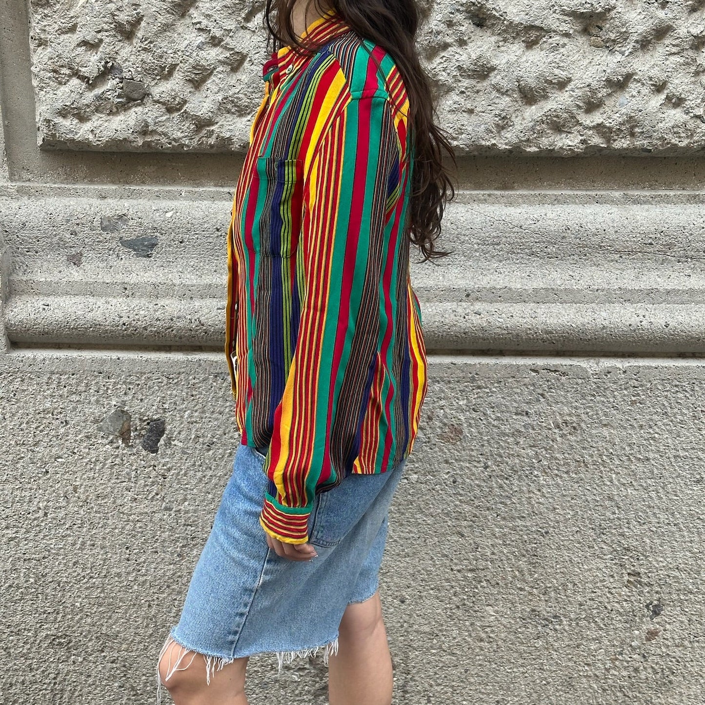 Person wearing a colorful striped shirt and denim shorts standing against a stone wall.