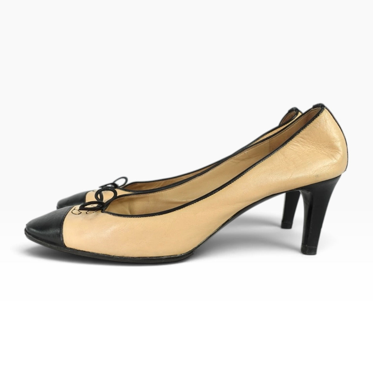 Chanel beige leather pumps with black cap toes, piping, and bow detail with CC logo on a mid-height heel.