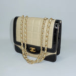 Side view of a vintage Chanel black and cream quilted flap bag showcasing its structured silhouette, patent leather details, and gold-tone chain straps.